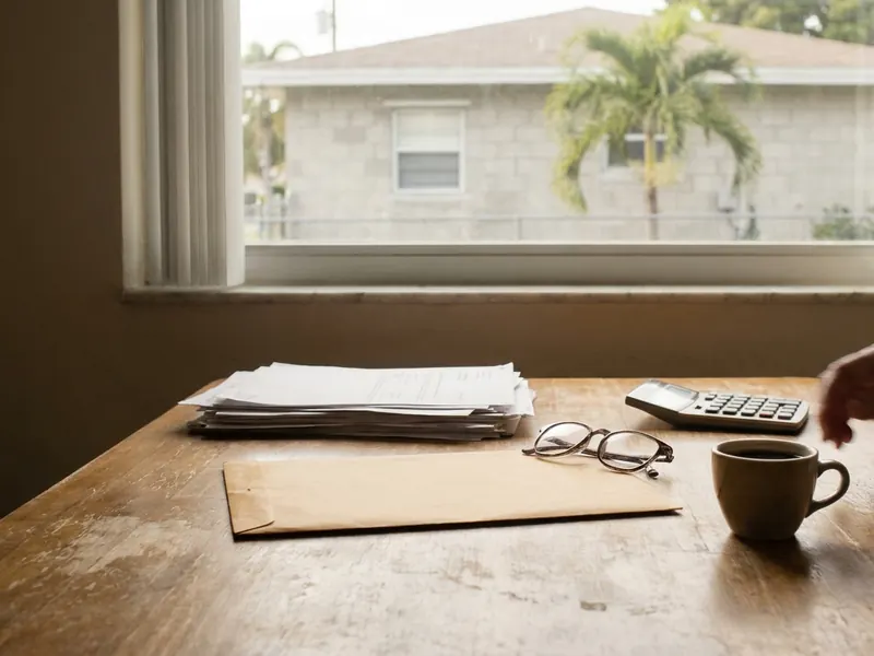 Kitchen table in a Miami home with an insurance renewal letter, envelope, reading glasses, calculator and coffee, a palm tree outside the window.